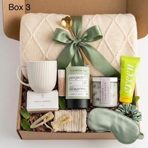 Könnte beinhalten: Eine Geschenkbox mit der Aufschrift Box 3 enthält eine cremefarbene Strickdecke mit salbeigrünem Band und goldenem Löffel, einen weißen gerippten Becher, Katili Sabun Seife, Grapefruit-Lavendel-Lippenbalsam, Tea With Tae Grüntee, Lavendel-Peeling und Schlafmaske.