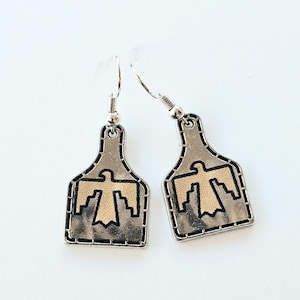 Pode incluir: Um par de brincos prateados com um desenho de pássaro-trovão dourado. Os brincos têm formato retangular com topo curvo e fecho de gancho. O pássaro-trovão é um símbolo de pássaro estilizado.