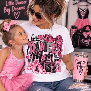 Può includere: Una t-shirt bianca con una stampa grafica che riporta il testo "DANCE MOM" in glitter rosa e argento, circondata da illustrazioni di una ballerina, un fiocco leopardato, scarpette da danza, una borsa sportiva e una borraccia con il testo "DANCE fuels my soul".