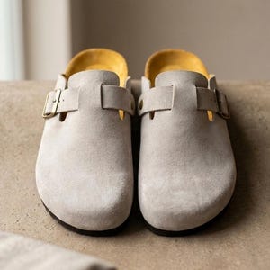 Puede incluir: Un par de zuecos de ante gris claro con punta redondeada, una tira con hebilla de metal dorado en el empeine y un forro interior de color amarillo brillante. Un pequeño logotipo circular con el texto Valero Leather aparece en la esquina inferior.