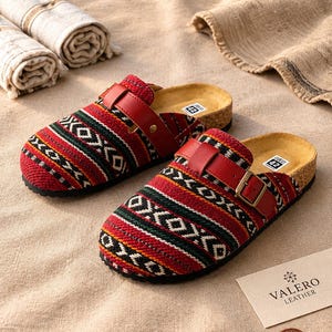 May include: A pair of red, green, and black patterned slip-on clogs with a red leather strap and metal buckle. The footbed is cork-colored with a size 42 label. A Valero Leather card and a circular wooden Valero Leather logo are present.