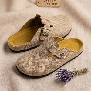 May include: A pair of beige hessian clogs with yellow insoles, black outsoles, and a metal buckle strap. A wooden tag labelled VALERO LEATHER is positioned above the shoes, and a small bundle of dried purple lavender is placed next to them.