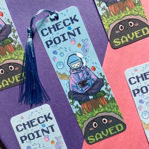May include: A light blue rectangular bookmark with a blue tassel, featuring a pixelated illustration of a person in a space helmet and hoodie playing a handheld game while sitting on a log, with the text CHECK POINT and SAVED in pixel font.