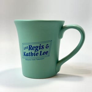 May include: A light green ceramic mug with a curved handle. The mug features the text "LIVE Regis & Kathie Lee" in blue, with the Buena Vista Television logo. The mug has a smooth, glossy finish and a slightly flared rim.