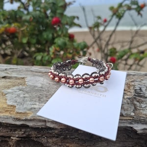 Könnte beinhalten: Ein Armband aus dunkelbraunem und weißem geknotetem Garn mit eingearbeiteten pfirsichfarbenen Perlen, das auf einer weißen Karte mit dem in Gold gedruckten Text AETERNEITH HAND-KNITTED LUXURY liegt und filigrane Spitzenmuster zeigt.