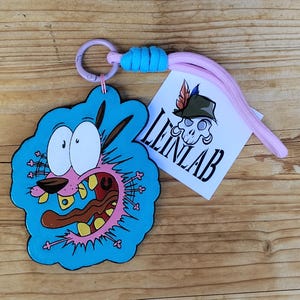 Può includere: Un portachiavi con un cane rosa dei cartoni animati con occhi spalancati e bocca aperta su sfondo blu, attaccato a un anello di plastica viola chiaro con un cordino blu brillante. Un'etichetta bianca quadrata mostra un logo con teschio e il testo LEINLAB.