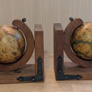 May include: A pair of bookends featuring two vintage-style globes mounted on dark brown wooden bases. Each globe is held by a curved wooden frame and accented with black metal corner brackets that feature visible rivets on the rectangular base.