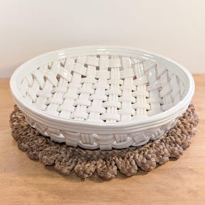 May include: A round, white ceramic bowl featuring a decorative woven basket-weave pattern throughout its structure. The bowl has a smooth, glossy finish and a raised, solid rim, showcasing an intricate lattice design that forms the base and sides.