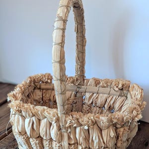Peut inclure: Un panier rectangulaire fabriqué à partir d'épis et de feuilles de maïs séchés. La base et les côtés présentent des épis de maïs verticaux, tandis que le bord supérieur est orné de feuilles de maïs séchées et froncées. Une anse arquée est entourée de fibres fines.
