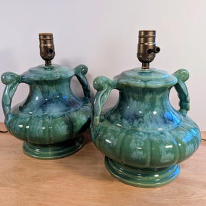 Peut inclure: Une paire de lampes de table avec une base en céramique en forme d'urne, dotée d'un vernis brillant aux nuances bleu sarcelle et vert. Chaque lampe présente deux anses décoratives, un corps cannelé et une douille métallique foncée.