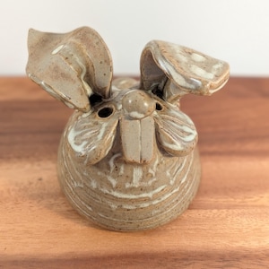 May include: A tan ceramic figurine shaped like a rabbit with a speckled glaze finish. The object features two prominent ears, a small round nose, and two rectangular front teeth. The surface has a textured, earthy appearance with white highlights.