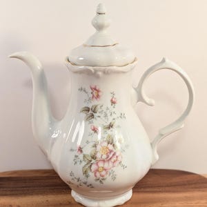 May include: A white porcelain teapot featuring a delicate floral pattern of pink roses and green leaves on the front. The teapot has a curved handle, a long spout, and a removable lid with a finial, all accented with thin gold trim.