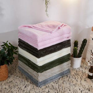 May include: A stack of six folded blankets featuring a textured waffle-knit pattern. The blankets are arranged in a color gradient from top to bottom, including shades of light pink, dark charcoal, cream, olive green, light gray, and blue-gray.