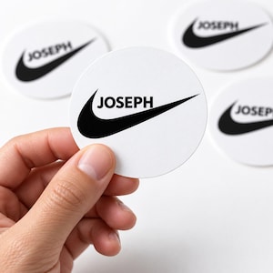 May include: A circular white sticker featuring a black swoosh logo and the name JOSEPH printed in black capital letters. The sticker is held in place, with three additional identical stickers visible in the background on a plain white surface.