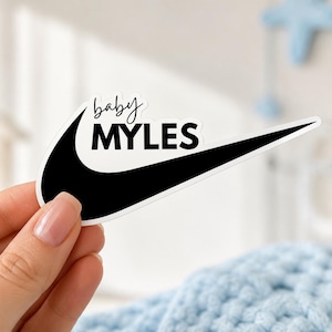 May include: A black and white sticker featuring a stylized swoosh shape with the text baby MYLES printed in black. The word baby is written in a cursive script font above the word MYLES, which is displayed in a bold, uppercase sans-serif font.