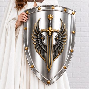 Könnte beinhalten: Ein großer, silberfarbener Wappenschild mit einem zentralen Relief eines Schwertes, das von dunklen, gefiederten Flügeln mit goldfarbenen Akzenten flankiert wird. Der Rand ist mit gleichmäßig verteilten goldfarbenen Nieten verziert.