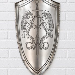 Könnte beinhalten: Ein silberfarbener Metallschild mit spitzem Ende und einem verstärkten Rand, der mit gleichmäßig verteilten Nieten versehen ist. In der Mitte des Schildes befindet sich ein schwarzes Gravurdesign eines vertikalen Schwerts.