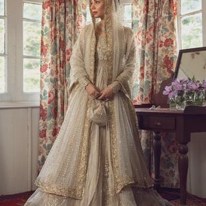 May include: A floor-length cream-colored gown featuring intricate gold embroidery and beadwork. The ensemble includes a matching sheer dupatta draped over the head and shoulders, long sleeves, and a small, coordinating drawstring pouch bag.