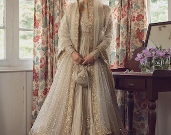 May include: A floor-length cream-colored gown featuring intricate gold embroidery and beadwork. The ensemble includes a matching long-sleeved bodice, a voluminous skirt, a sheer dupatta draped over the head, and a small matching drawstring pouch.