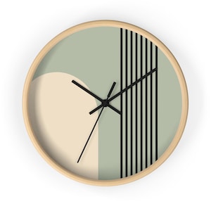 Peut inclure: Une horloge murale ronde avec un cadre en bois clair, présentant un fond vert sauge, un grand demi-cercle crème sur la gauche et une série de lignes verticales noires sur la droite. L'horloge possède trois aiguilles noires au centre.