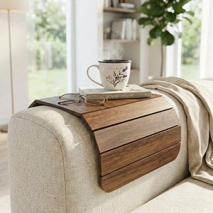 May include: A flexible, dark brown wooden sofa arm tray with a slatted design that drapes over the armrest. It holds a white ceramic mug with a botanical print, a book titled Scandi Design, and a pair of thin-rimmed reading glasses on top.