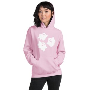 May include: A light pink hooded sweatshirt featuring a front kangaroo pocket and a graphic print of three white ghosts with simple facial expressions centred on the chest. The garment has long sleeves and a matching drawstring hood.