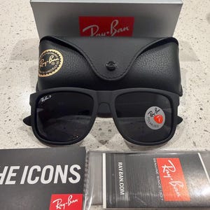 May include: Matte black Ray-Ban sunglasses with polarized lenses, featuring a black leather-textured carrying case, a silver branded box, and two informational booklets with the text THE ICONS and RAY-BAN.COM, GENUINE SINCE 1937.