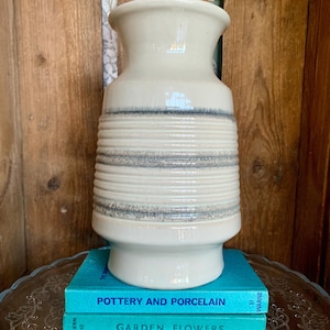May include: A cream-coloured ceramic vase with horizontal textured bands and a flared top. The vase sits atop two teal books titled "Pottery and Porcelain" and "Garden Flowers". The background is a wooden wall.