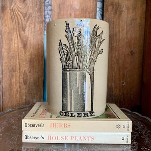 Może przedstawiać: Ceramiczny kubek z czarno-białym rysunkiem łodyg selera w słoiku. Kubek ma nadrukowane słowo "CELERY". Kubek stoi na trzech książkach zatytułowanych "Observer's HERBS", "Observer's HOUSE PLANTS" i "WARNE 85".