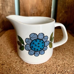 May include: White ceramic creamer with a blue and green floral design. The flower has a blue center with white stars and is surrounded by green leaves.