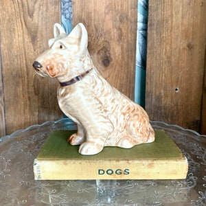 May include: A ceramic figurine of a brown and white Scottish Terrier dog sitting on a book titled "Dogs".
