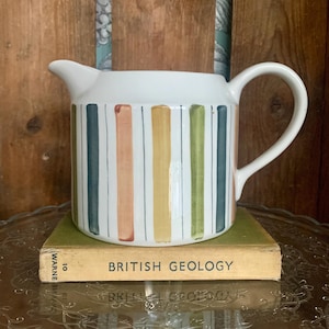 Op de afbeelding: Witte keramische kan met een kleurrijk gestreept ontwerp in tinten groen, blauw, geel en roze. De kan heeft een handvat en staat op een boek met de titel "British Geology".