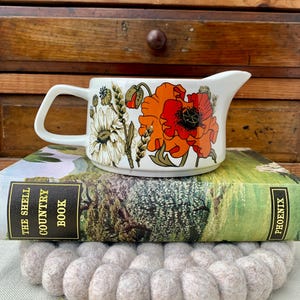 May include: A white ceramic creamer with a handle and spout, decorated with vibrant orange and red poppy flowers and other floral elements. The creamer sits atop a vintage book and a gray felt trivet, against a wooden background.