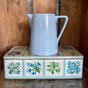 以下が含まれることがあります： 取っ手付きの薄い青色のセラミックピッチャーが、花柄のヴィンテージなブリキ箱の上に置かれています。箱には、ブルーベル、勿忘草、その他の野花など、さまざまな花柄の4つのパネルがあります。