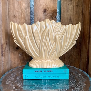 Pode incluir: Um vaso de cerâmica amarelo claro com um design de folhas estilizado. O vaso está em cima de dois livros com os títulos "Plantas de interior" e "Flores de jardim".