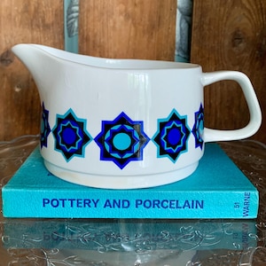 May include: White ceramic gravy boat with a blue and black geometric pattern. The pattern features a repeating design of stars with a turquoise center.