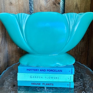 May include: A large, turquoise ceramic vase with a flared, petal-like design. The vase sits atop a stack of three books with titles including "Pottery and Porcelain," "Garden Flowers," and "House Plants."