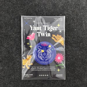 Puede incluir: Un paquete rectangular de plástico que contiene un pin circular azul con ilustraciones blancas de tigres gemelos y escritura tailandesa. El empaque presenta el texto Yant Tiger Twin, Yant Suea Koo y gráficos decorativos de tigres y flores.