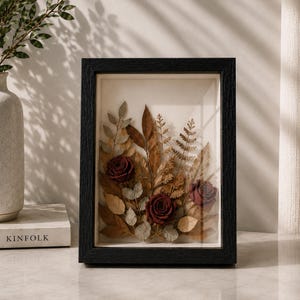 Peut inclure: Un cadre rectangulaire noir contenant un arrangement floral pressé sur un fond crème. La composition présente trois roses rouge foncé entourées de diverses feuilles séchées brunes et vert pâle ainsi que des frondes de fougère sous verre.