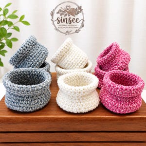Peut inclure: Trois ensembles de deux paniers au crochet empilés en bleu, crème et magenta. Chaque panier présente une construction en fil épais texturé avec une forme arrondie et une large ouverture. Le logo sinsee artisanat au crochet & créations faites main est visible.