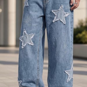 May include: Light blue denim jeans featuring a wide-leg silhouette and multiple star-shaped cutouts with frayed, distressed edges. The denim has a classic medium wash finish, visible stitching along the seams, and a standard button and zipper fly.