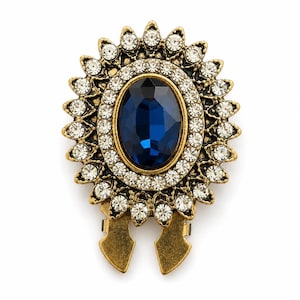 Puede incluir: Un broche decorativo con una gran piedra central ovalada de color azul oscuro, rodeada por dos anillos concéntricos de pequeños diamantes de imitación blancos transparentes en un marco de metal dorado antiguo con un clip de dos puntas en la base.