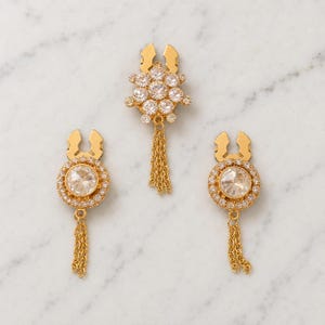 Op de afbeelding: Een set van drie goudkleurige sieraden met heldere, gefacetteerde kristallen. Het middelste stuk is een cluster van kristallen met een bungelende kettingfranje, terwijl de twee zijstukken ronde kristallen knopjes met kettingen zijn.