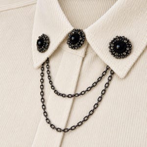 Pode incluir: Uma camisa canelada na cor creme com uma gola adornada por três broches circulares. Cada broche possui um cabochão preto central rodeado por pequenos cristais escuros. Duas correntes de metal escuro conectam os broches, pendendo à frente.