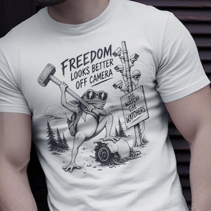 May include: A white short-sleeved t-shirt featuring a black graphic of a frog wearing sunglasses and holding a sledgehammer. The text reads FREEDOM LOOKS BETTER OFF CAMERA and WATCH THE WATCHERS, accompanied by an illustration of security cameras.