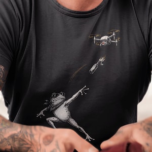 Pode incluir: Uma t-shirt preta de manga curta com uma estampa gráfica de um sapo usando óculos de sol e uma bandana, atirando uma garrafa em chamas em direção a um drone. O sapo está representado numa pose dinâmica com um braço estendido.