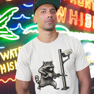 Może przedstawiać: Biały t-shirt z czarno-białą grafiką przedstawiającą szopa pracza używającego piły szablastej do przecinania metalowego słupa, na którym zamontowana jest kamera bezpieczeństwa z napisem FLOCK. Wzór znajduje się na środku klatki piersiowej.