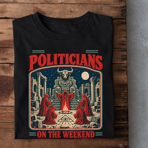 May include: A black t-shirt featuring a graphic print of four figures in red robes gathered around a fire in front of a stone bull statue and ancient ruins. The text "POLITICIANS" appears above the image and "ON THE WEEKEND" appears below it.