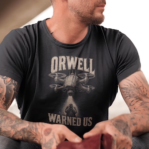 May include: A black short-sleeved t-shirt featuring a graphic print of a drone hovering above a person. The text ORWELL is printed above the drone, and the text WARNED US is printed below the person, with the drone casting a spotlight downward.