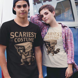 Peut inclure: Un t-shirt noir et un t-shirt crème, chacun arborant un graphique de raton laveur portant une boîte en carton sur la tête. Le texte "SCARIEST COSTUME" est imprimé au-dessus du raton laveur et le mot "FLOCK" figure sur le côté de la boîte.